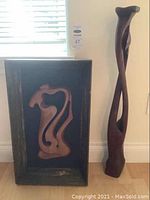 Photo showing two wood sculptures; one tall twisting carved wood sculpture next to the framed wood sculpture with abstract carved figure inside a rectangular wooden frame.