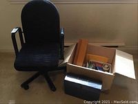 Photo of the office chair together with the box of office supplies and black document organizer on a beige carpeted floor.