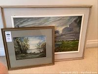 Two framed landscape paintings; smaller watercolor painting in foreground with gold frame, larger acrylic landscape painting behind with wide beige frame.