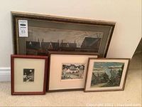 Wide shot showing four framed artworks arranged on carpeted floor against wall, various sizes, depicting buildings and urban scenes.