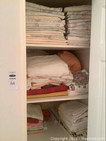 Closet shelf with neatly folded bedding, blankets, and tablecloths in pastel and light colors with floral patterns.