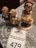 Two ceramic Hummel figurines displayed on a lace tablecloth, one with a seated girl and goose, another figurine of boy and girl standing.