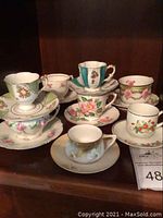 Group photo showing nine assorted porcelain teacups and saucers arranged on a shelf, various floral patterns and colors visible.