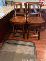 Pair of wooden barstools shown together near countertop with three foam kitchen mats placed on wooden floor in front.