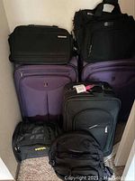 Photo showing full collection of luggage and carry-on bags stacked together, including two large purple suitcases, one black carry-on suitcase, and three smaller black carry-on bags.