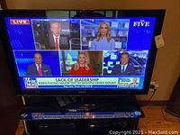 Samsung 50-inch TV powered on, showing news channel screen with multiple guests in dialogue. The TV is set on a stand with remote controls visible underneath.