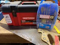 Closed Craftsman toolbox, Ryobi drill accessory set plastic case, and hand saw with wooden handle on a wooden surface.