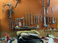 Wide view of a variety of metal hand tools including wrenches, sockets, hanging on a pegboard above a cluttered workbench.