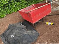Photo showing red wooden wheelbarrow with two handles and one wheel, along with black grill cover and mat partly visible.