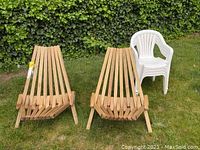 Two wooden slatted lawn chairs next to three stacked white plastic lawn chairs on grass with ivy backdrop
