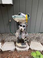 Front view of outdoor garden bear yard art holding a fishing rod with a large fish on its head, placed on concrete blocks surrounded by gravel and soil.