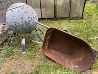Photo of spherical garden composter with ventilation holes and green metal legs, alongside a rusted metal wheelbarrow on grass.