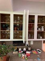 Wooden kitchen cabinets filled with various glasses and mugs, including clear wine glasses, colored stemmed glasses, and plastic mugs.