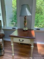 Front view of Ethan Allen end table with lamp on top near windows, showing wood top and cream painted base with drawer.