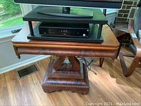 Full view of vintage wooden pedestal table under TV with hinged top and wheels.