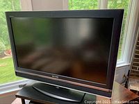 Front view of Sony 32 inch television on black oval stand beside a window with a green outdoor scene visible.