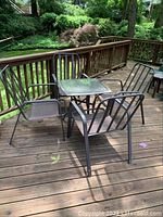 Outdoor metal table with glass top and four matching metal arm chairs, positioned on wooden deck with surrounding greenery.