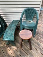 Photo shows three outdoor furniture pieces: a curved wooden bench with peeling paint, a round wooden stool table, and a single green plastic chair.