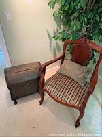 Photo showing wood framed armchair featuring curved arms, striped cushioned seat, green decorative pillow, and caster wheels standing next to the metal covered trunk.