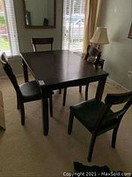 Full view of wooden dining table with 4 matching chairs around it, placed in a room with window and lamp nearby. Chairs have black faux leather seats.