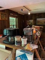 Two coffee makers side by side with ceramic crocks and other small kitchen items on table with wood panel walls background