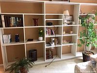 Wide shot showing white shelving filled with books, faux plants, decorative shells, a milk glass lamp, and clocks.