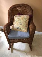 Front view of brown plastic wicker rocking chair with blue seat cushion and two throw pillows (floral with ruffle trim and solid burgundy).