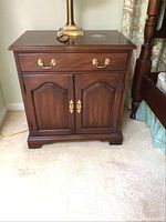 Front view of one wooden nightstand showing top surface, single drawer with brass handles, and double cabinet doors with brass hinges and handles.