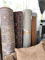 Three area rugs rolled and standing vertically, showing one patterned rug with reds and browns and one plush shag rug partially disclosed.