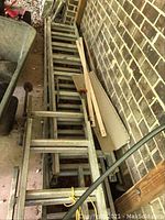 Two aluminum extension ladders stacked outdoors against a brick wall, showing an array of metal rungs and weathered condition.