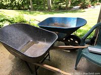 Two wheelbarrows shown outdoors on pavement, one black True Temper brand and one blue, both with wooden handles and single wheels, weathered condition.