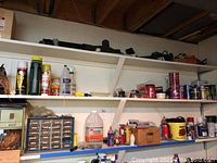 Wide view of white shelving loaded with assorted household items including pest and garden chemicals, paint cans, hardware organizers, and bottles.