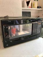 Black GE microwave oven on a white surface with reflective door showing some reflection of surroundings.