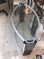 Oval glass top coffee table with metal frame and lower glass shelf viewed from above and side.