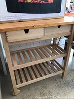 Front view of wooden kitchen cart showing drawer with dovetail joints and hooks underneath plus slatted shelf and rolling wheels.