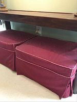 Pair of matching rectangular upholstered ottomans with burgundy slip covers featuring piping edges. Positioned side by side under a console table on carpeted floor.