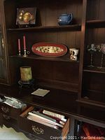 Wooden shelving and drawers holding red taper candles in silver holders, pillar candle on stand, fruit painting, ceramic blue teapot, ceramic platter, napkins and napkin rings, and rolled linens.