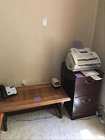 Photo showing the wooden file cabinet with two drawers and metal pulls, small wooden coffee table with legs, and Brother fax machine placed on top of the cabinet in a corner room