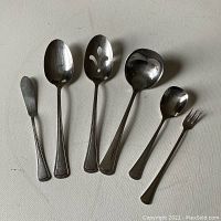 Full view of 6 utensils: butter knife, 2 serving spoons, ladle, small spoon, serving fork on a flat surface