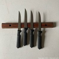 Wooden magnetic knife strip mounted on wall with 4 black-handled steak knives arranged vertically.