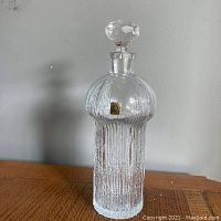 Full view of the clear crystal decanter with textured surface and faceted stopper, showing modernist form and label.