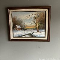 Framed winter scene painting hanging on gray wall showing full artwork with cabin, snow, trees, and sunset.