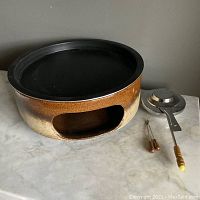 Side view of the ceramic Kera grill showing the fat lava glaze and the opening for the burner. The non-stick top plate is black and round. Adjacent to the grill are the mineral spirit burner and two wooden handled skewers used for cooking. The ceramic surface has a smooth texture with a gradient from brown to cream-yellow.