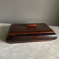 Closed view of rectangular wooden jewelry box with reddish wood and curved edges, sitting on marble surface