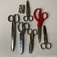 Six pairs of scissors arranged on a white surface including varying sizes and handle types.