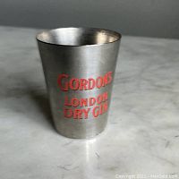 Photo of vintage metal shot glass with red 'GORDON'S LONDON DRY GIN' text on one side, placed on a white marble surface.