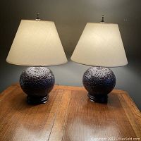Pair of purple-blue ceramic table lamps with round wooden bases, cream fabric shades, lit on a wooden surface.