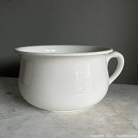 Side view of white ceramic antique chamber pot with handle on a marble surface.