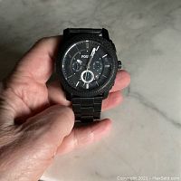 Front view of Fossil Machine Chronograph black stainless steel watch held in hand showing dial with three subdials, textured bezel, and white markers on black face.