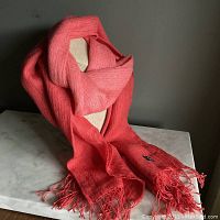 Red gradient scarf displayed draped over a beige mannequin bust, showing the full length and texture along with the fringe tassels at the end. The scarf is wound around the neck and laying down in folds on a marble surface.